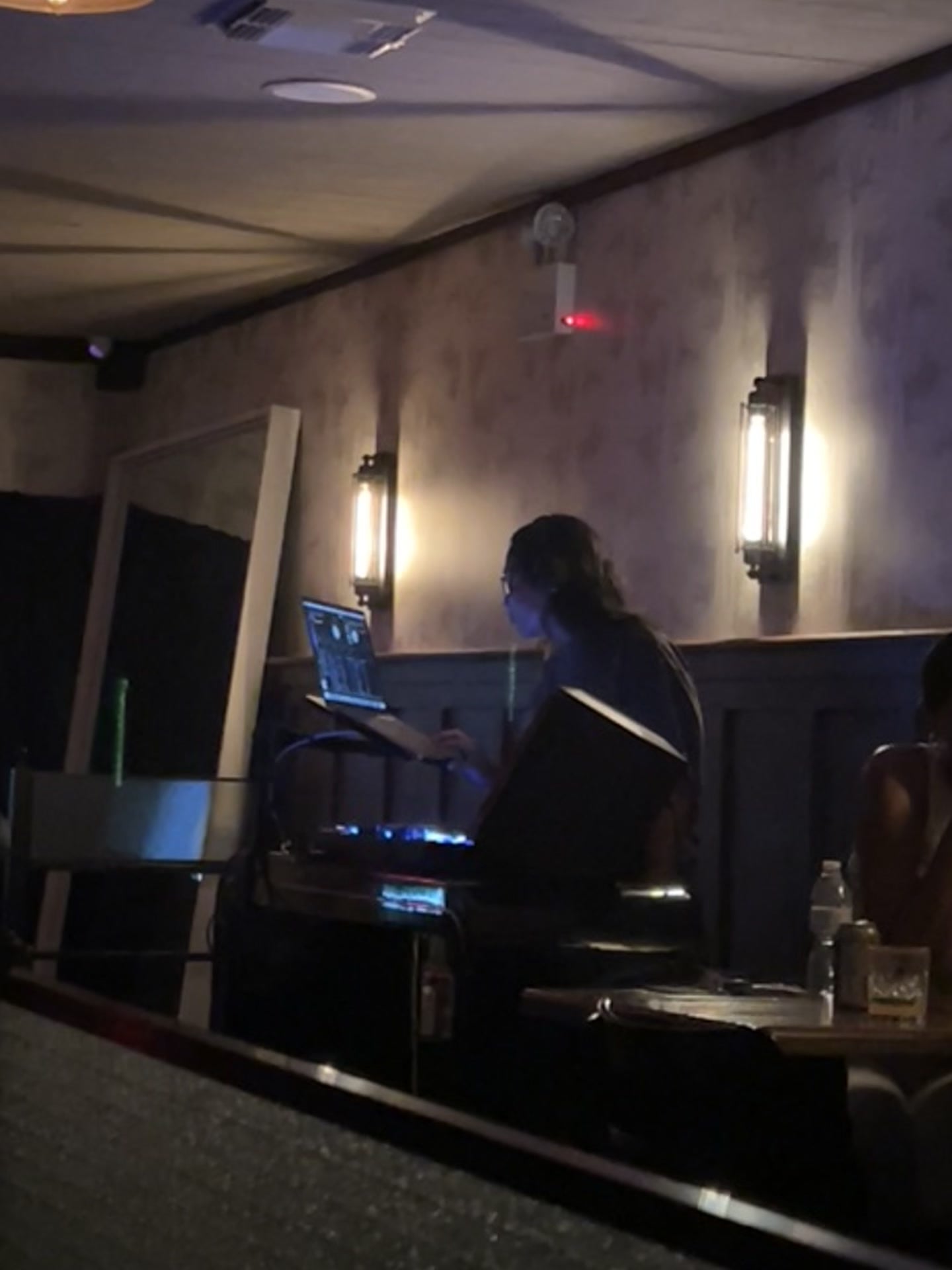 The Woodlot booth seen across the darkened room, La Phiesa in profile at her laptop between two glowing wall sconces.