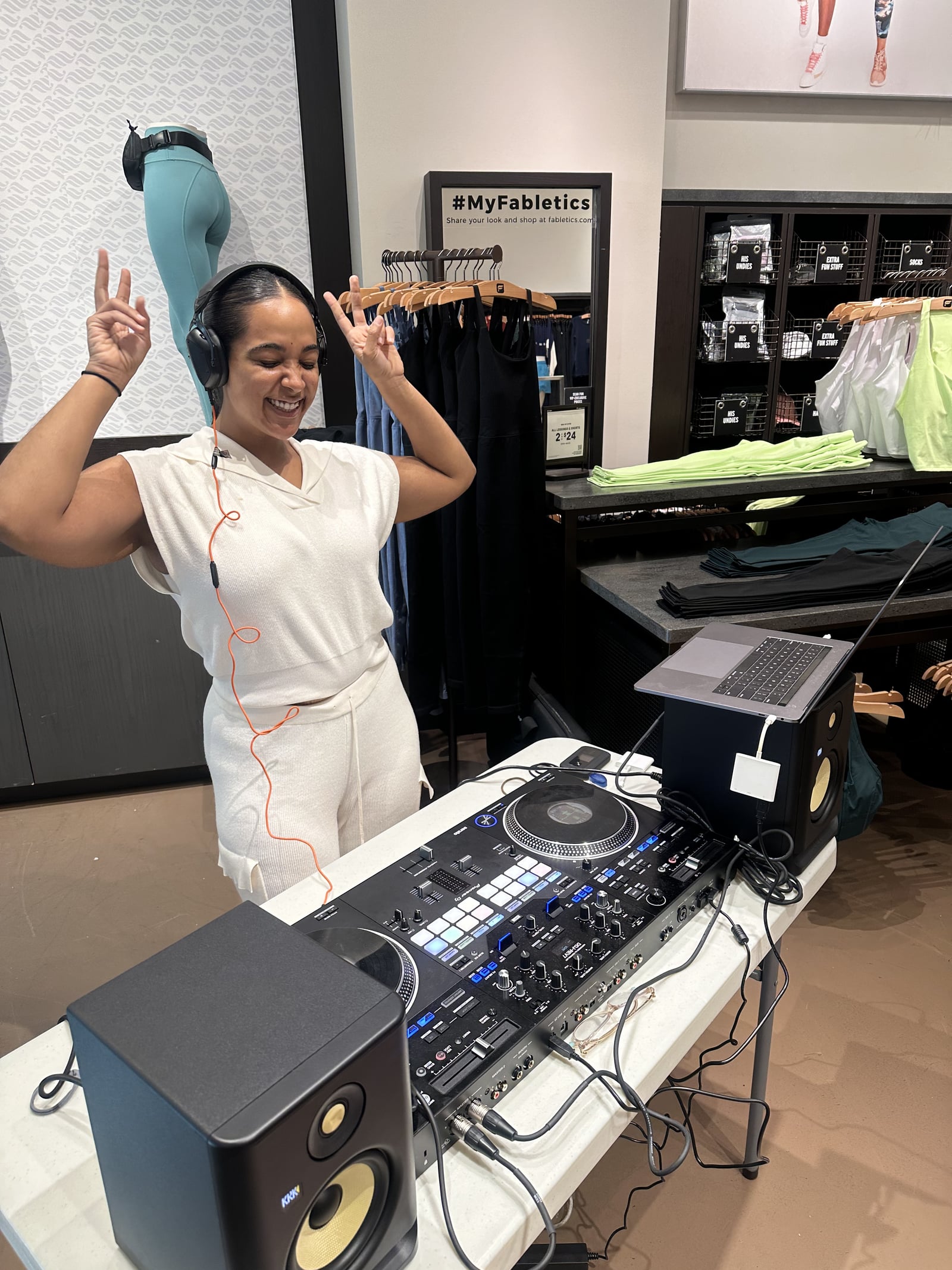 La Phiesa throwing both hands up mid-set at the Fabletics activation, the store's athleisure racks turned dance floor behind her.