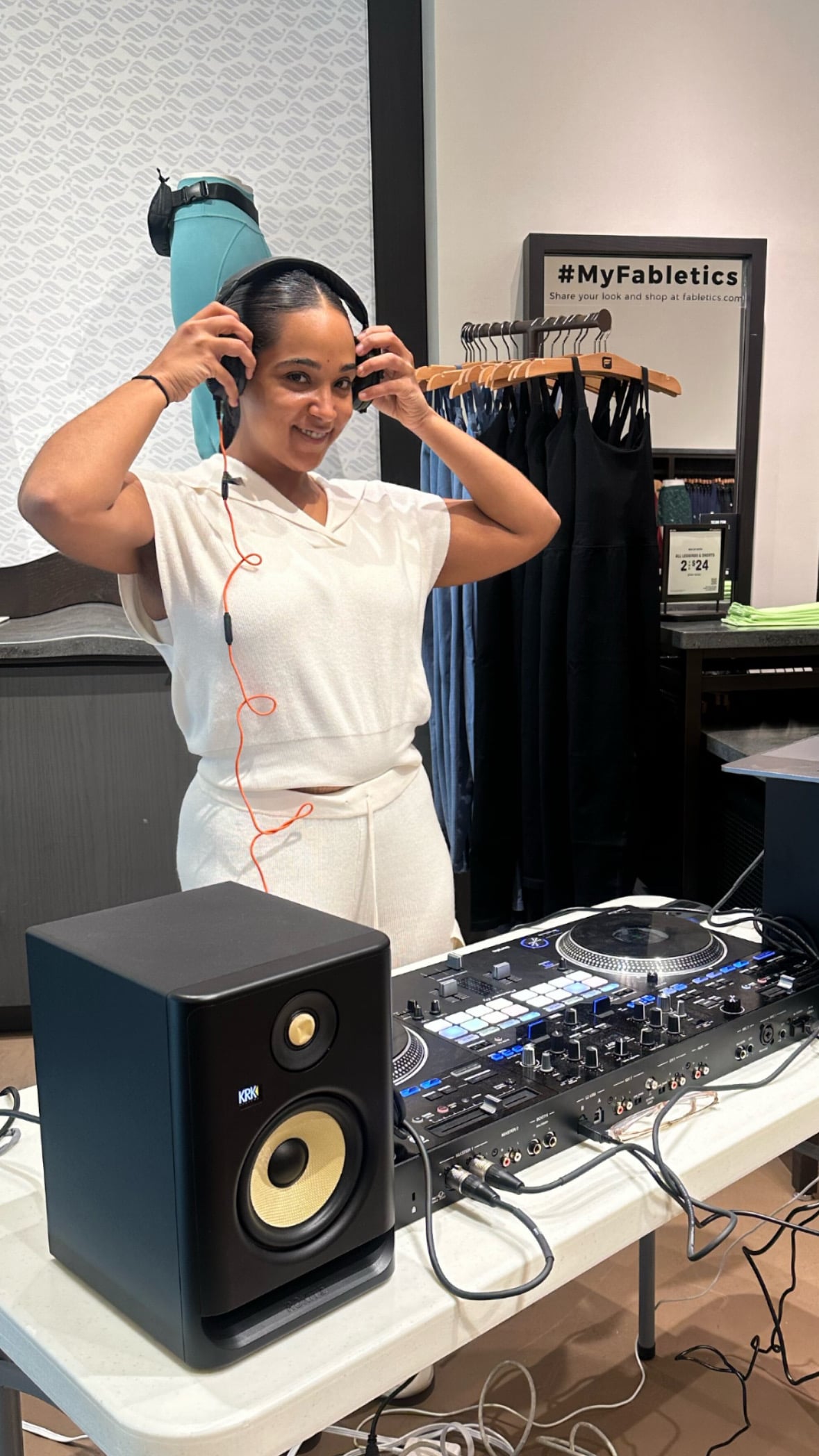 La Phiesa pulling her headphones on behind the decks at the Fabletics store event, the #MyFabletics board and legging racks framing the booth.