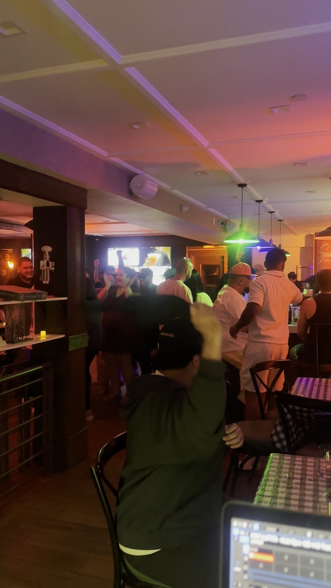 A packed room at Saucey seen from behind the decks, the crowd bunched under the bar's purple and green wash while La Phiesa's set runs.