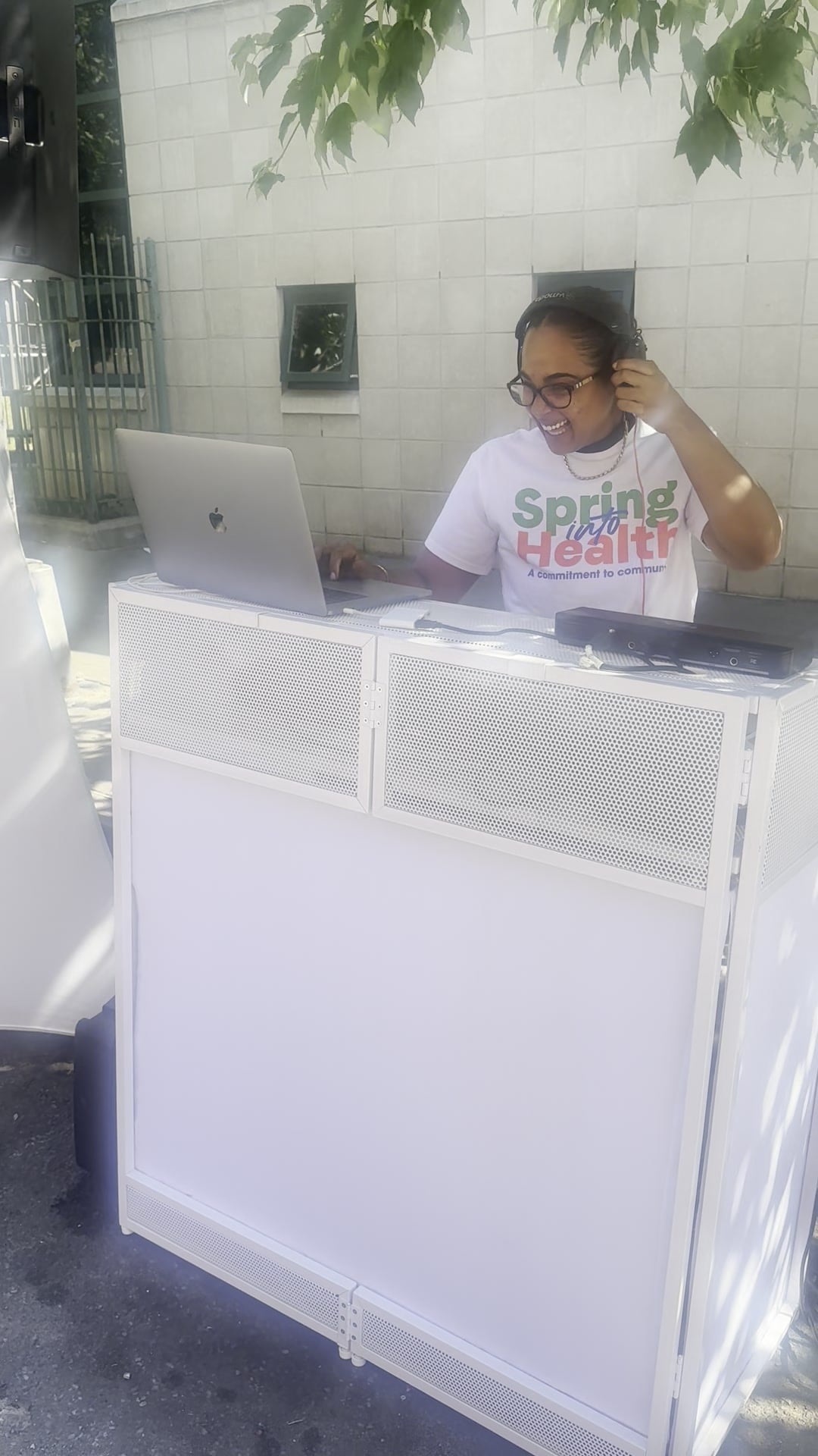 La Phiesa laughing behind the booth at the Spring into Health block party, event tee on and headphones in hand