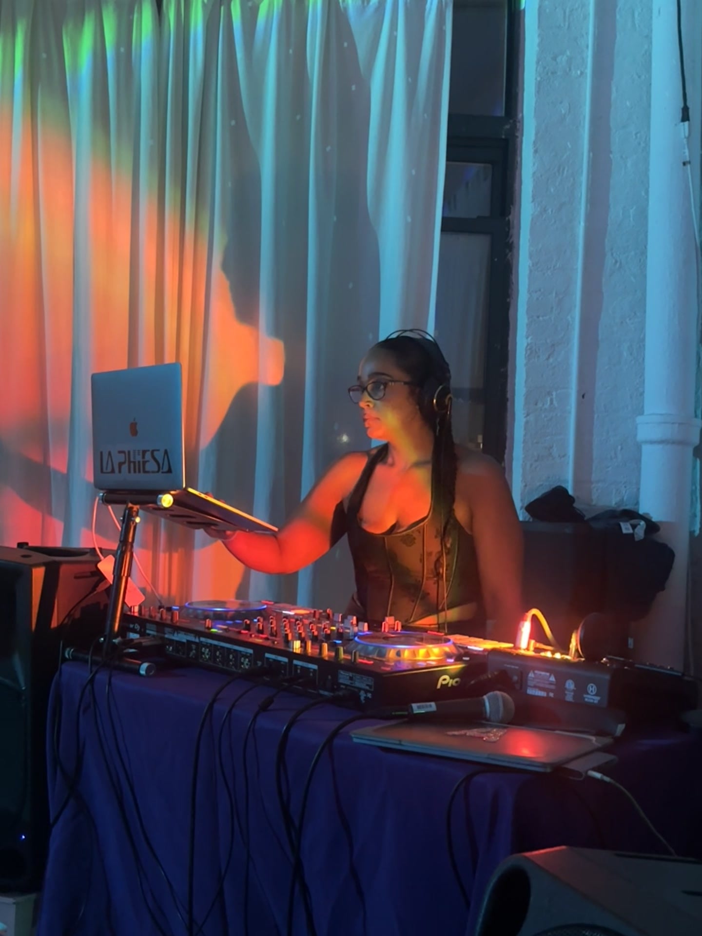 The full La Phiesa booth at Hussle Lab, branded laptop on its stand and Pioneer decks glowing against loft curtains washed in orange and teal.