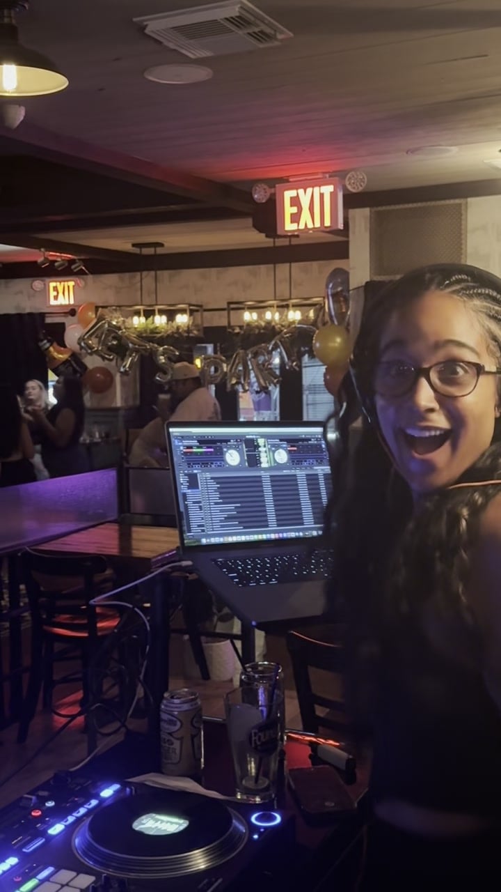 La Phiesa turns from the decks with a wide grin as balloons and a full room fill the frame behind her at Woodlot.