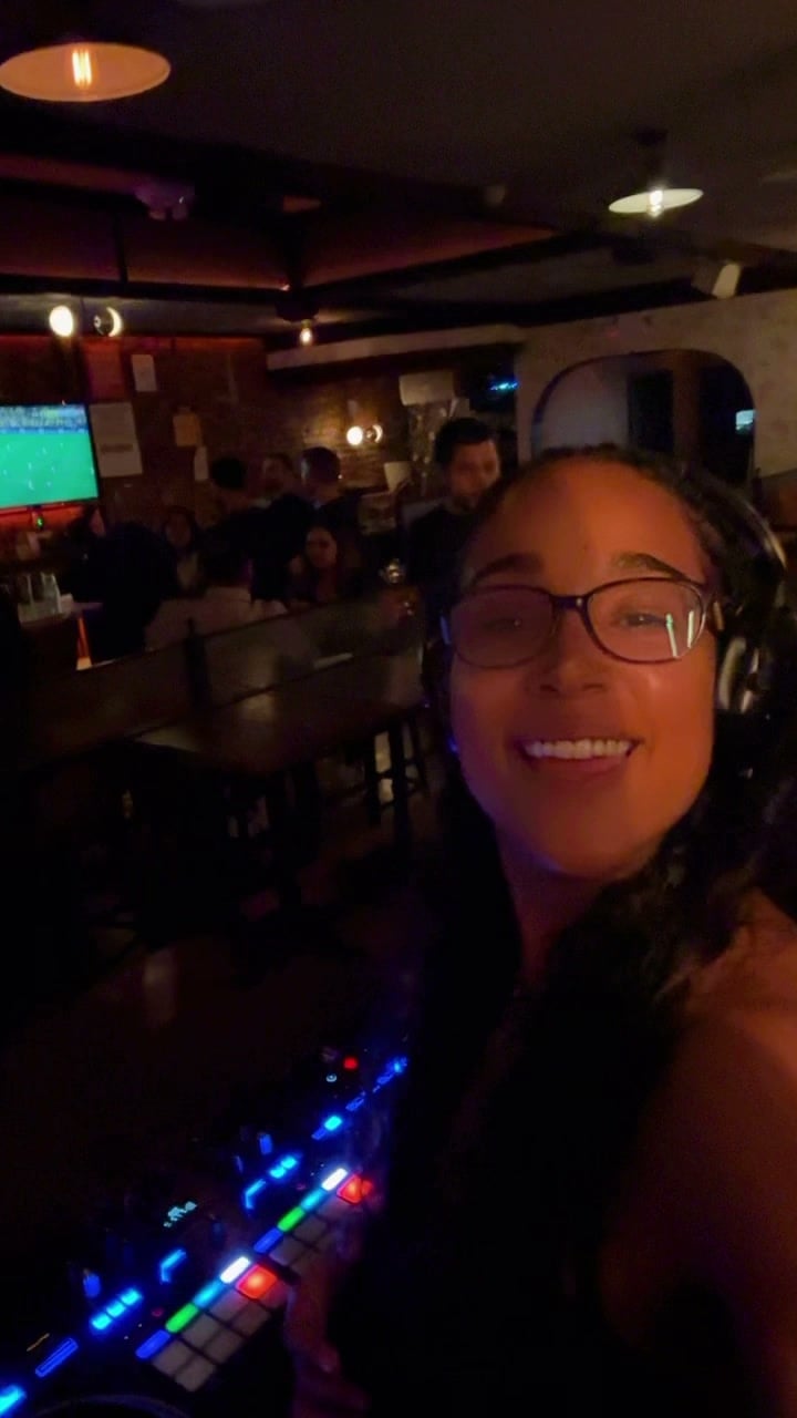 La Phiesa grinning with one headphone on at Woodlot in late June, the controller's coloured pads lit up in front of her.