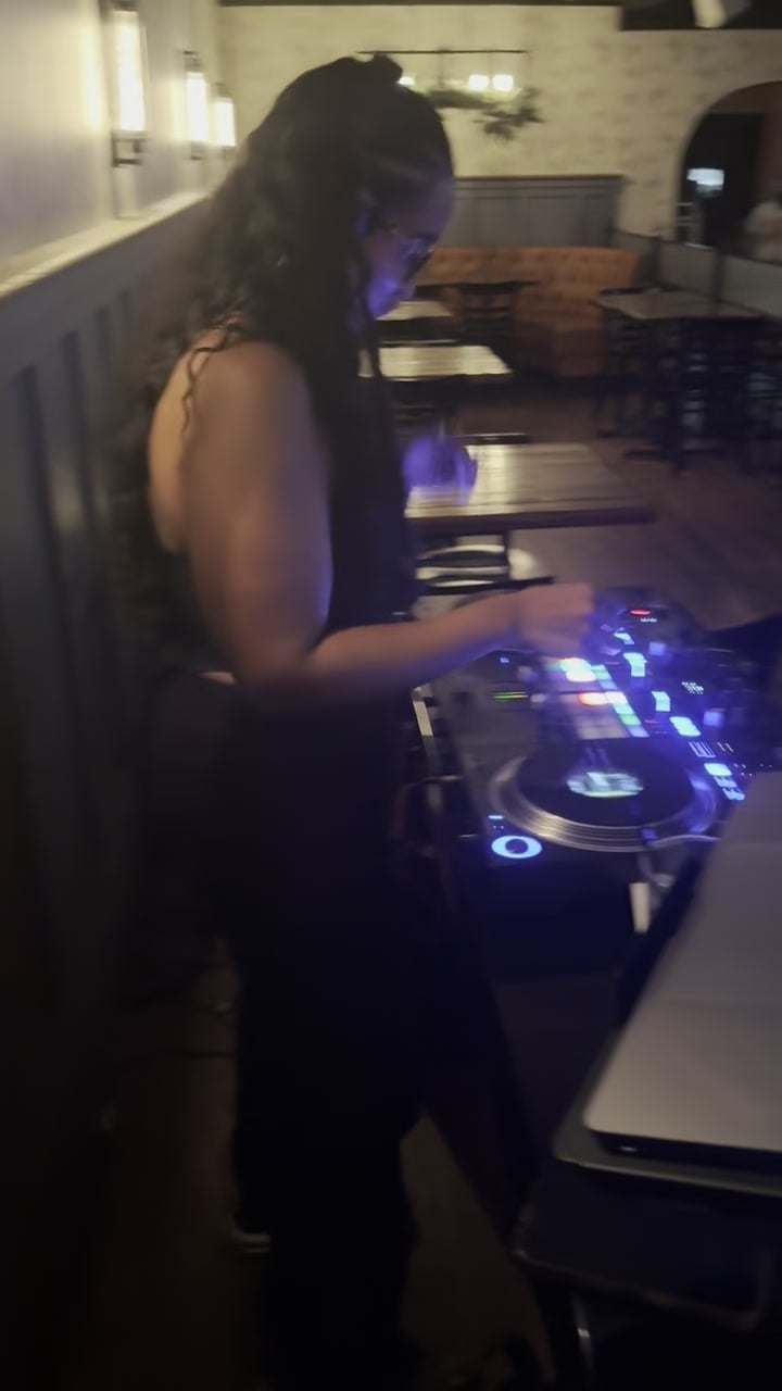 La Phiesa in profile with both hands on the CDJs at Woodlot, the June crowd's tables stretching out behind the booth.
