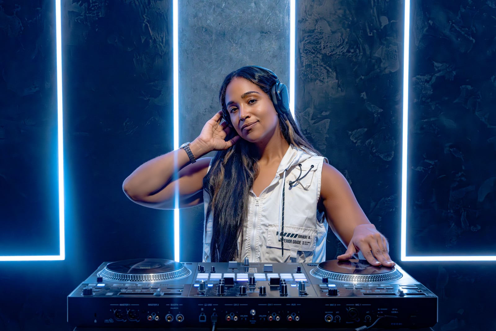 La Phiesa at the decks in a cream vest, hand to her headphones, blue neon strips